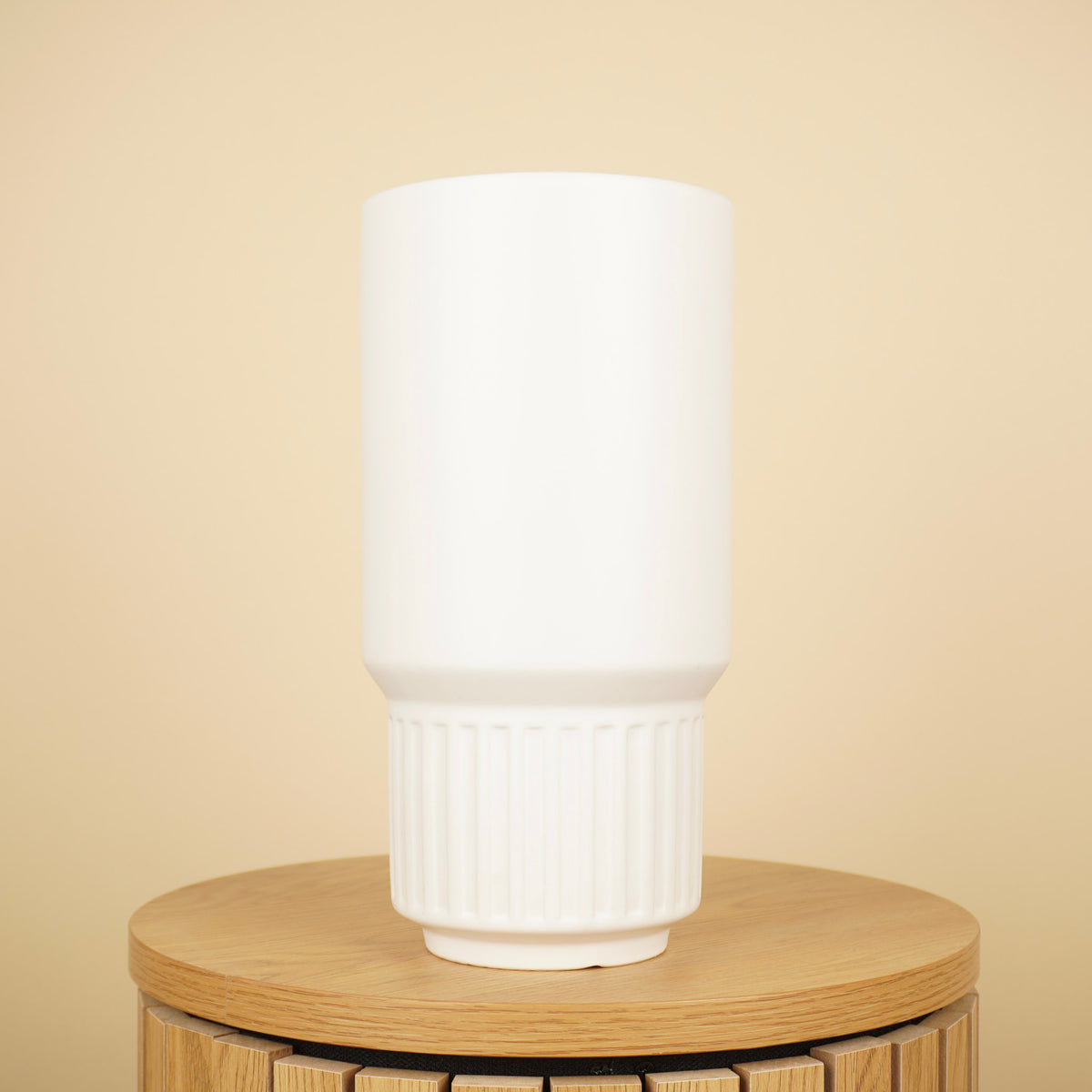 Hoge witte vaas met cilindervormige bovenzijde en geribbelde onderkant op een ronde houten tafel.