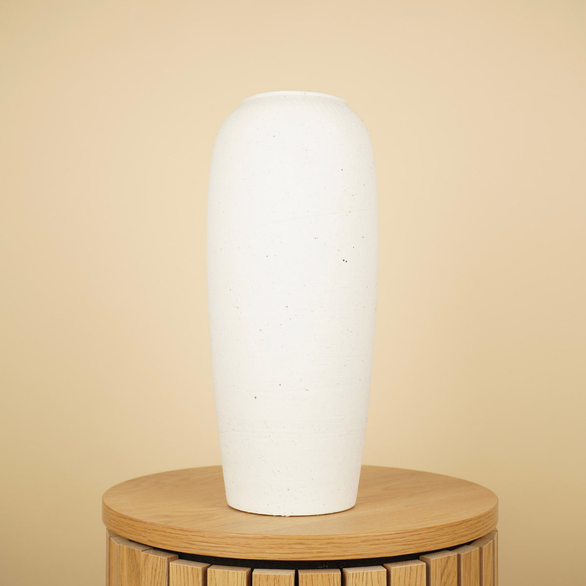 Hoge, slanke crèmekleurige vaas met matte afwerking en fijne spikkels op een ronde houten tafel.