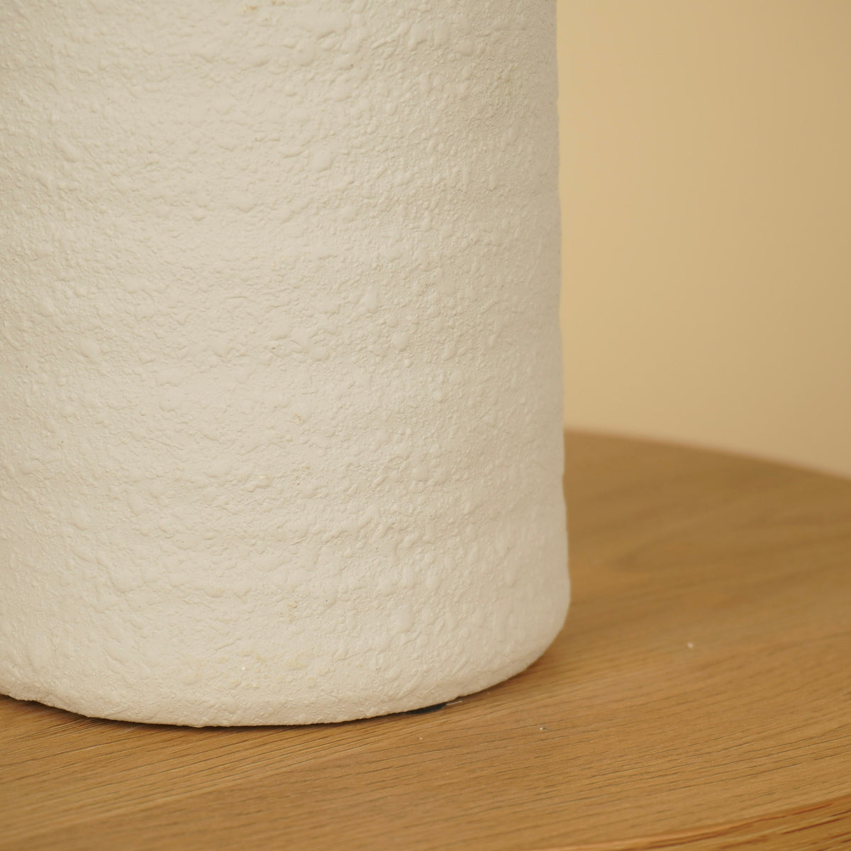 Detail van het ruwe, matte oppervlak en de afgeronde onderrand van een witte vaas op een houten tafel.