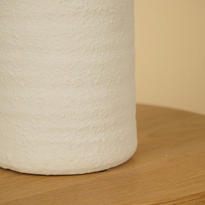 Detail van het ruwe, matte oppervlak en de afgeronde onderrand van een witte vaas op een houten tafel.