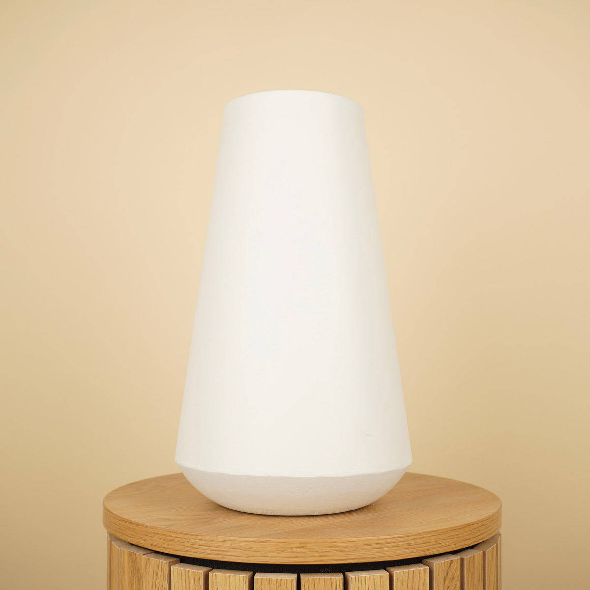 Hoge matwitte vaas met taps toelopende vorm en afgeronde beige voet op een ronde houten tafel.